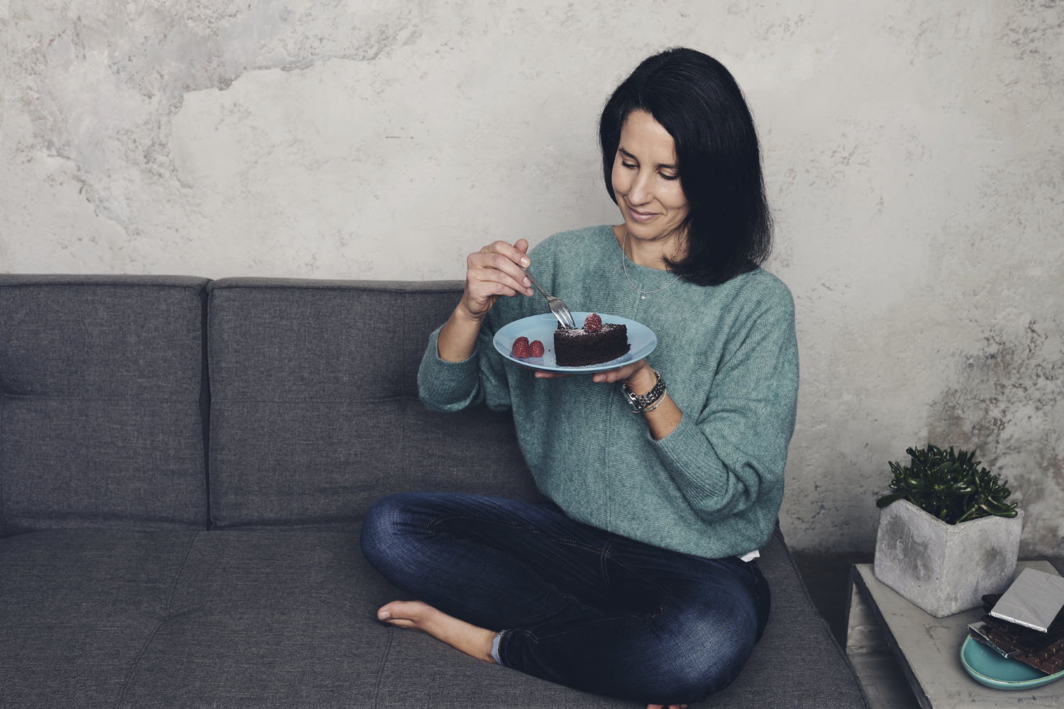 Barbara Plaschka sitzt auf dem Sofa, einen Teller mit Schokoladenkuchen vor sich und freut sich auf den nächsten Bissen. Genießerisch zum Wohlfühlgewicht